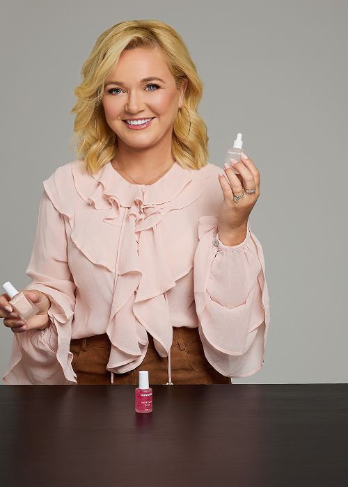 A smiling blonde woman in a pink ruffled blouse holds bottles of nail care products.