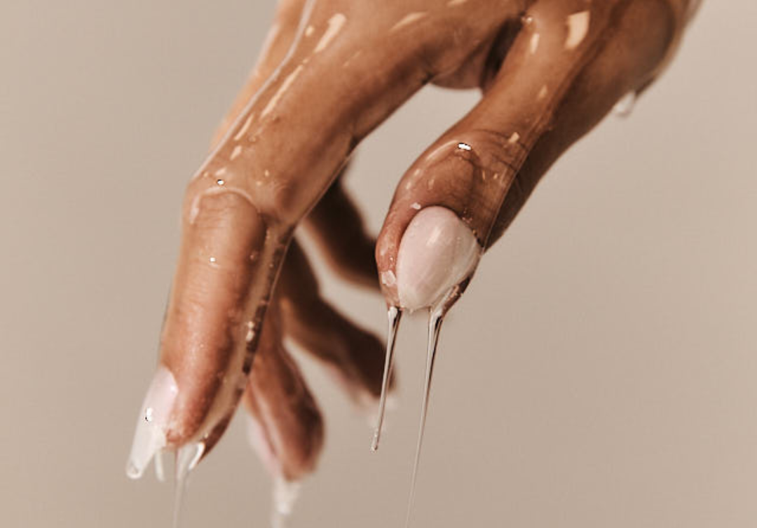 A close-up of a manicured hand covered in a clear, viscous liquid dripping from the fingertips.