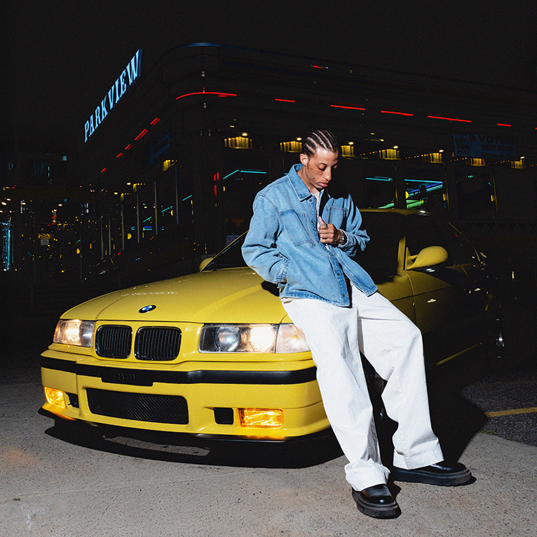 Person in denim jacket leaning on yellow car at night.