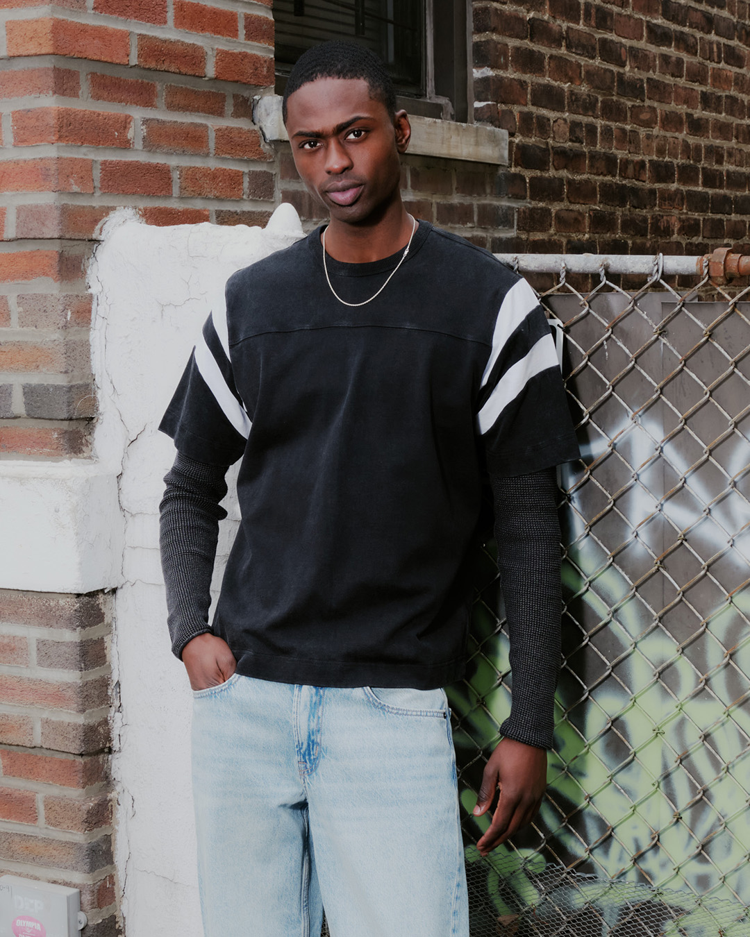 A man in a black striped t-shirt and jeans leans against a brick wall and a chain-link fence.