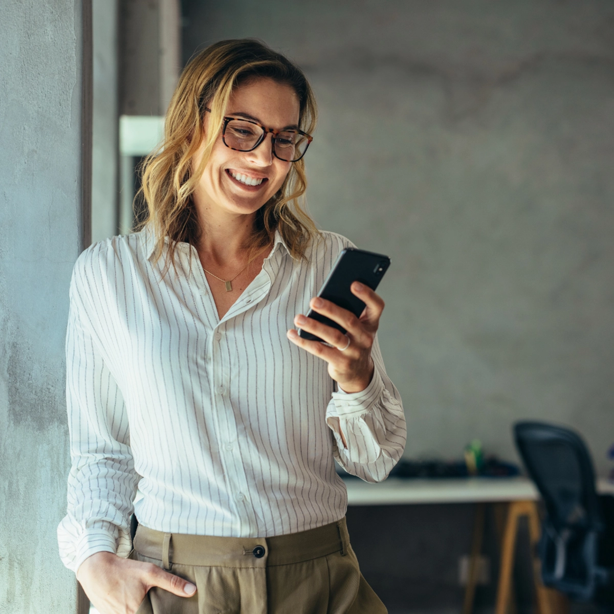 Person smiling while looking at a phone, standing indoors against a wall.