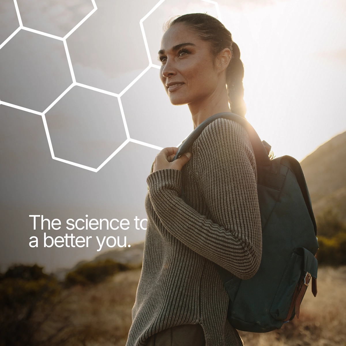 A woman with a backpack smiles as she stands on a hill with the sun shining behind her.