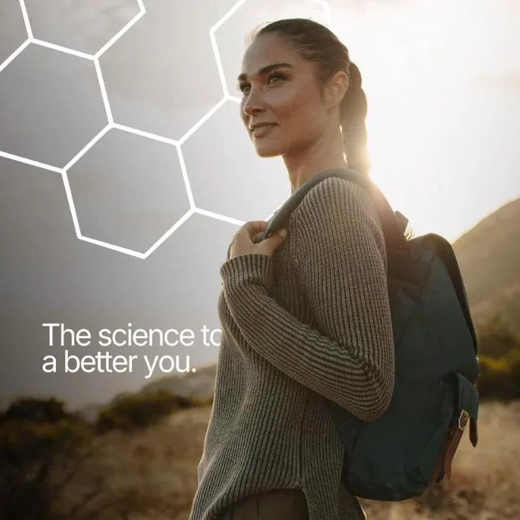 A woman with a backpack smiles as she stands on a hill with the sun shining behind her.