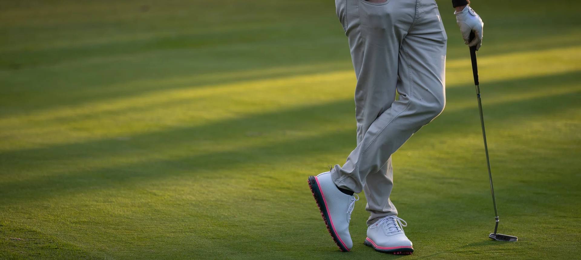 Golf player holding putter, standing on green grass, wearing white shoes.