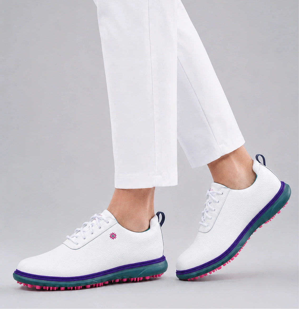 A person wears white trousers and white golf shoes with purple, teal, and pink soles.