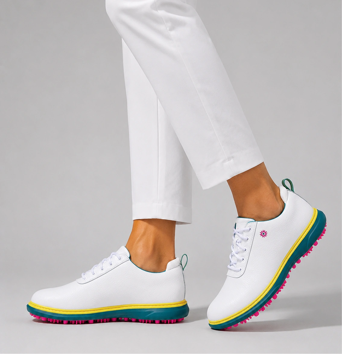 A person wearing white pants and white golf shoes with teal, yellow, and pink soles.