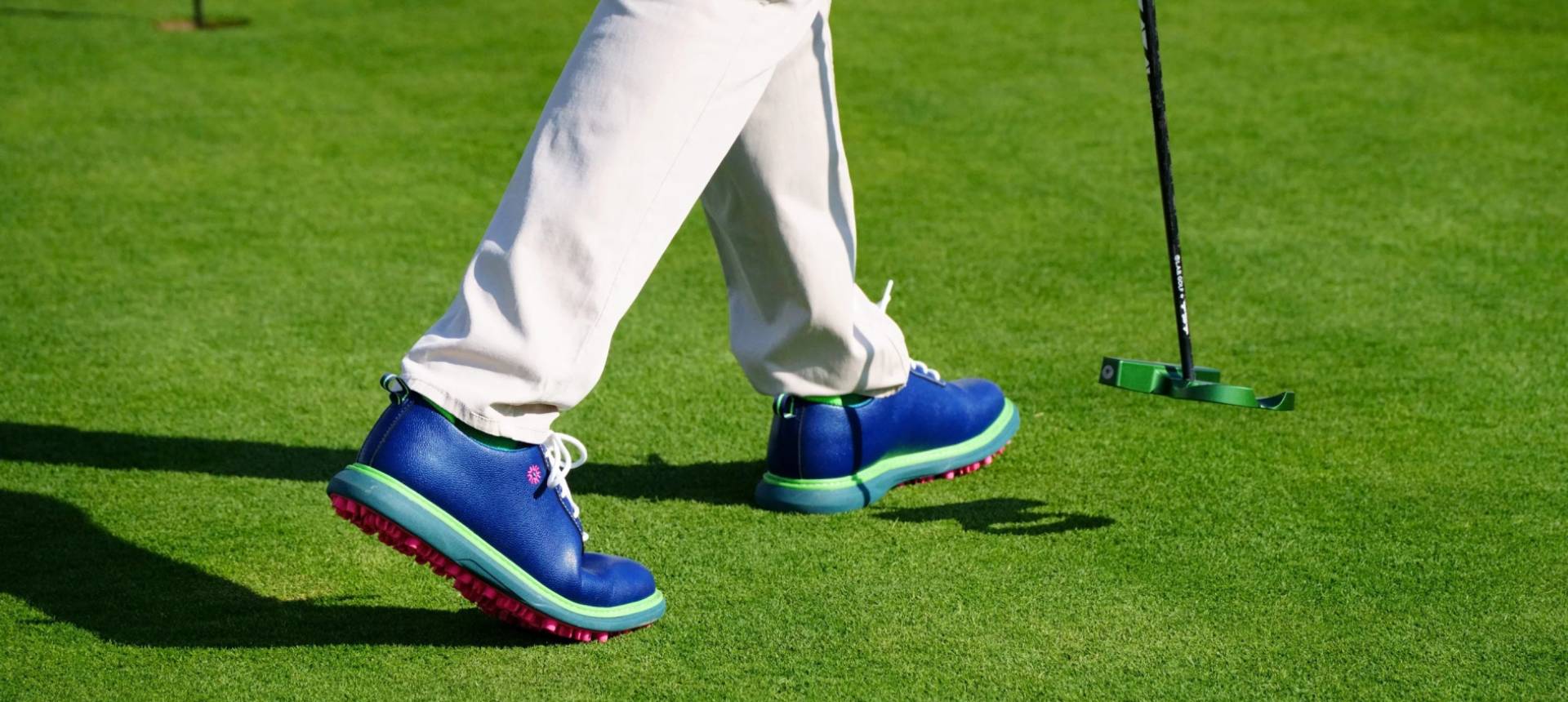 Person in blue shoes and white pants holding a green putter on grass.
