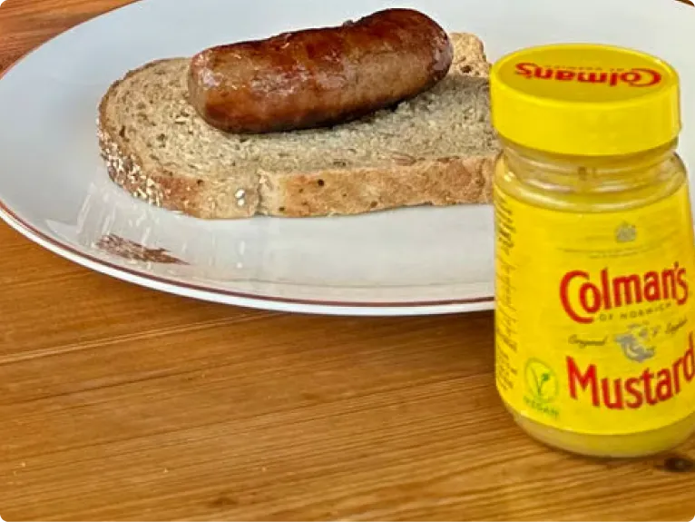 A cooked sausage on a slice of seeded bread on a white plate, next to a jar of Colman's mustard.