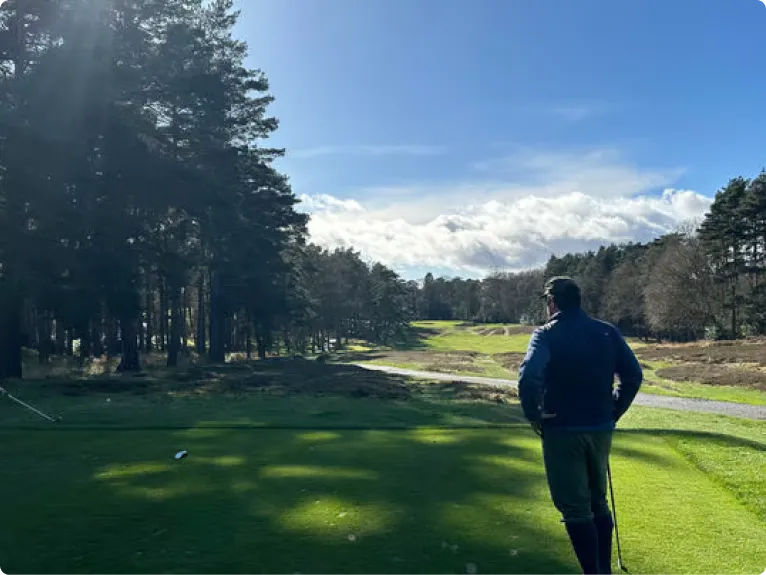 A man seen from behind on a golf tee box, looking down a sunny, tree-lined fairway.