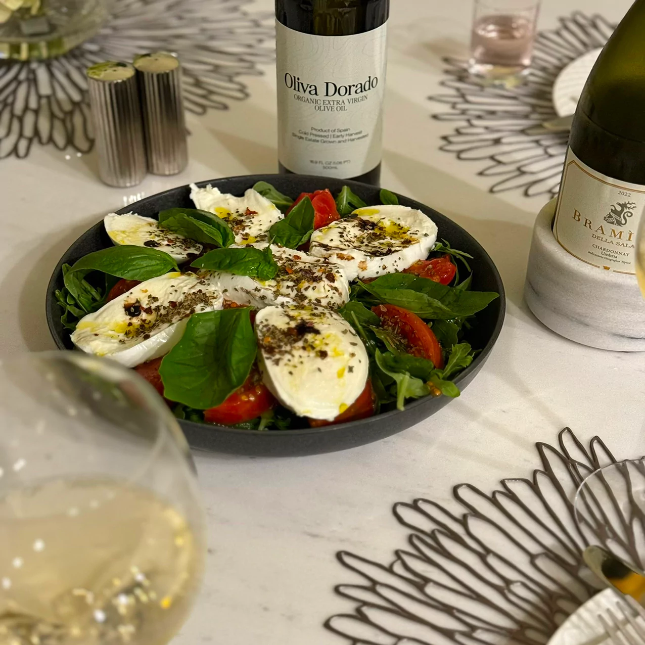Caprese salad with mozzarella, tomatoes, basil, and olive oil on a table with wine bottles.