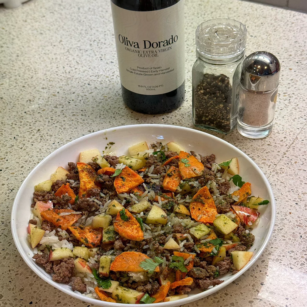 Bowl of mixed rice, vegetables, and meat with olive oil, pepper, and salt on a countertop.