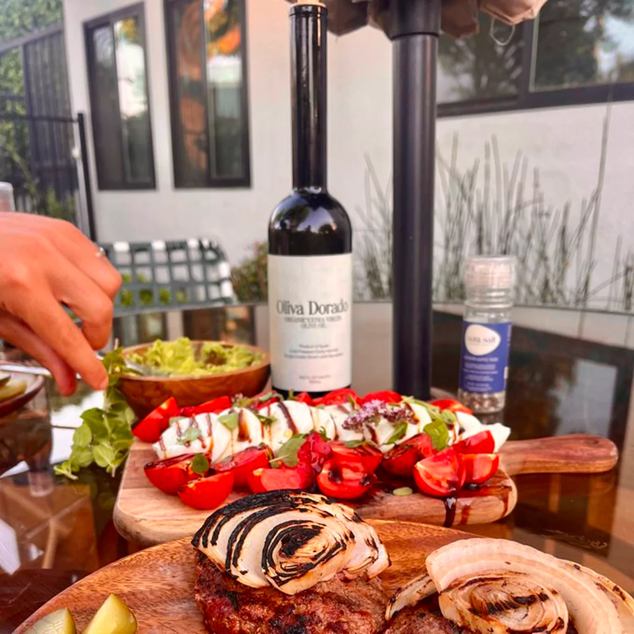 Outdoor table with grilled onions, salad, olive oil bottle, and a hand reaching for food.
