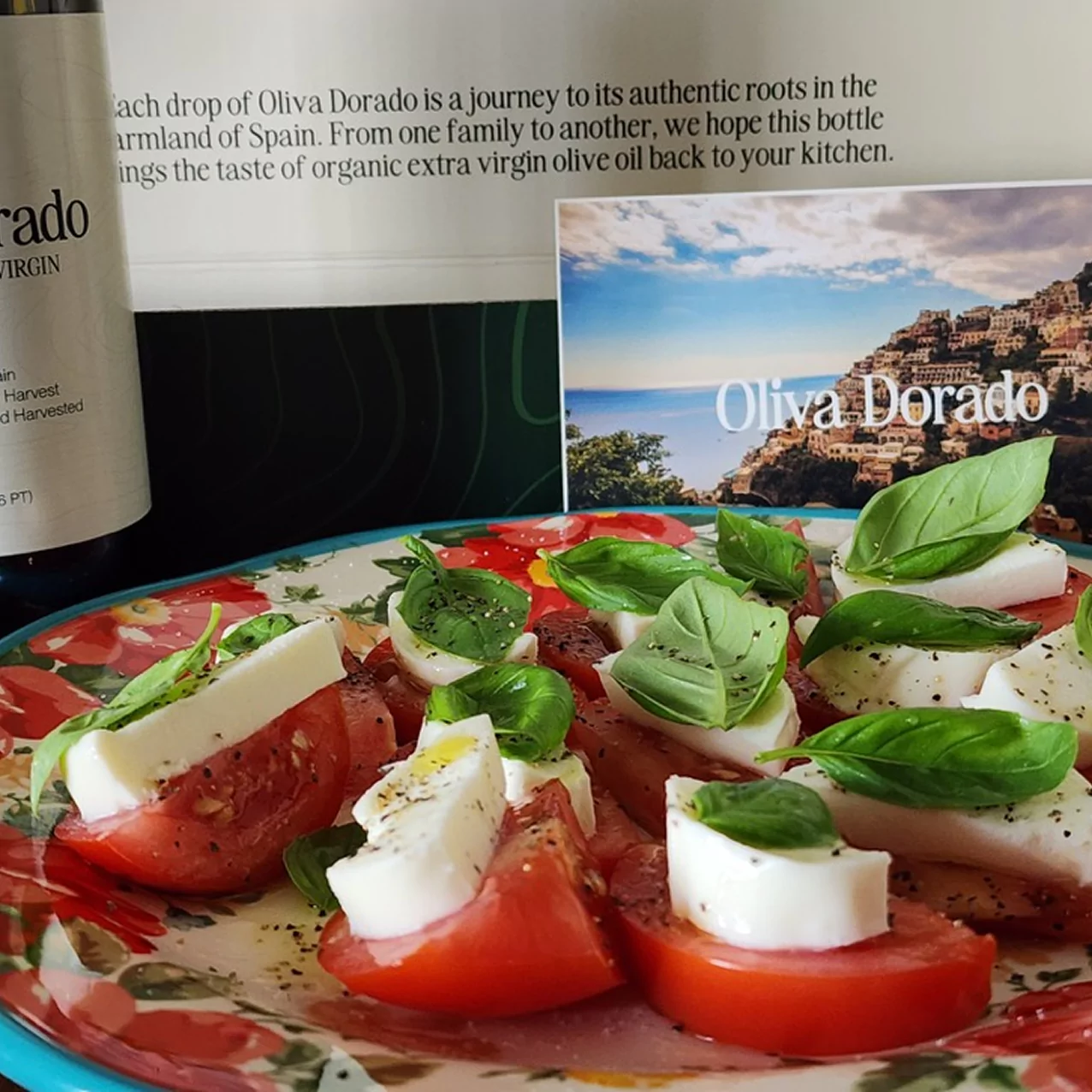 Caprese salad with tomatoes, mozzarella, and basil on a colorful plate, with olive oil bottle and postcard.