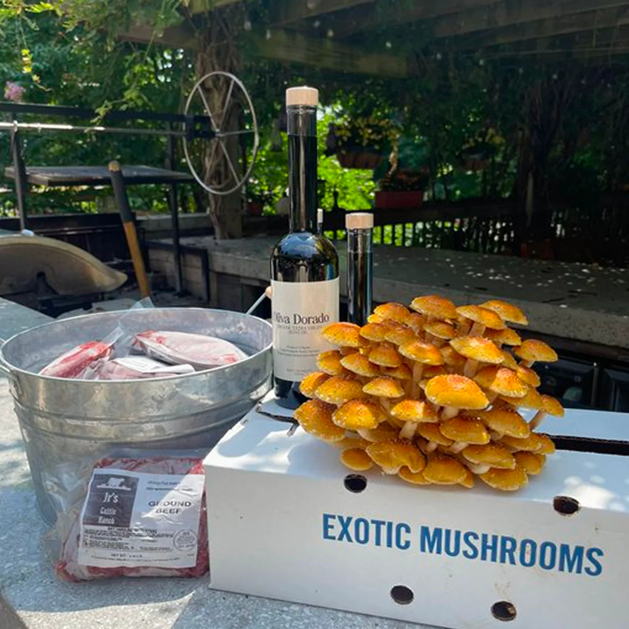 Box of exotic mushrooms, wine bottle, and meat on an outdoor table.