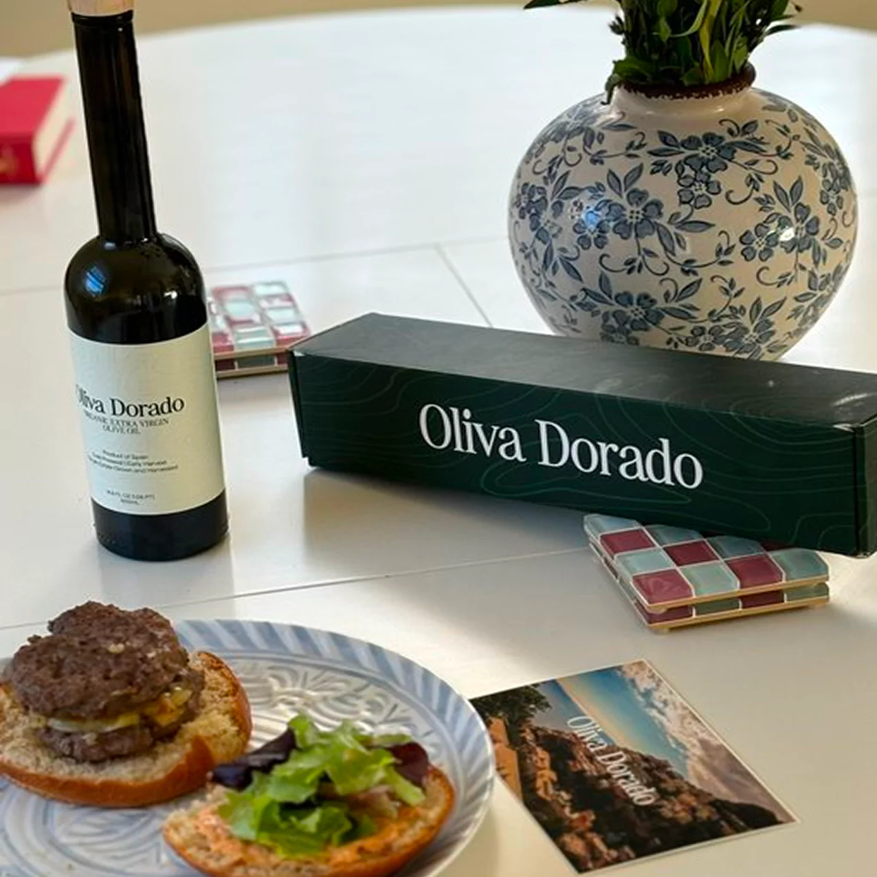 Bottle of olive oil, burger, and decorative vase on a table.