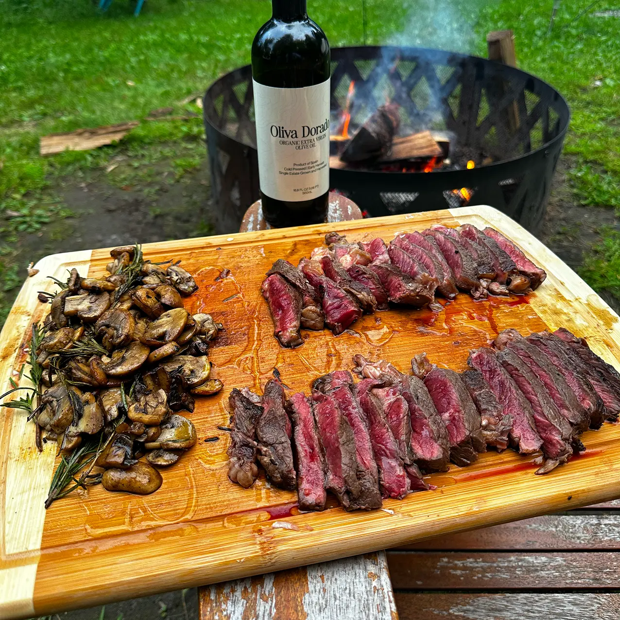 Sliced steak and mushrooms on a wooden board with a wine bottle and fire pit in the background.