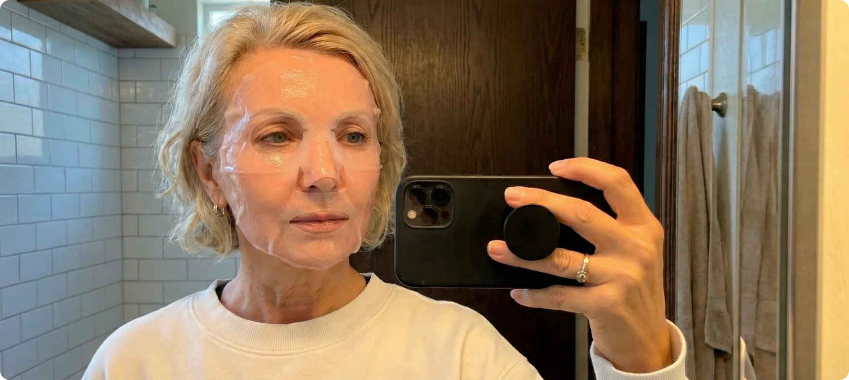 A woman wearing a clear sheet mask takes a selfie in a bathroom mirror.
