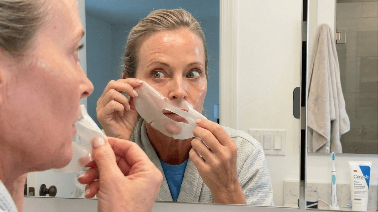 Person in bathroom mirror applying a sheet face mask.