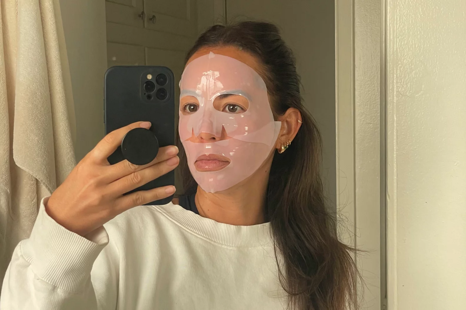 Person taking a selfie wearing a sheet mask in a bathroom.