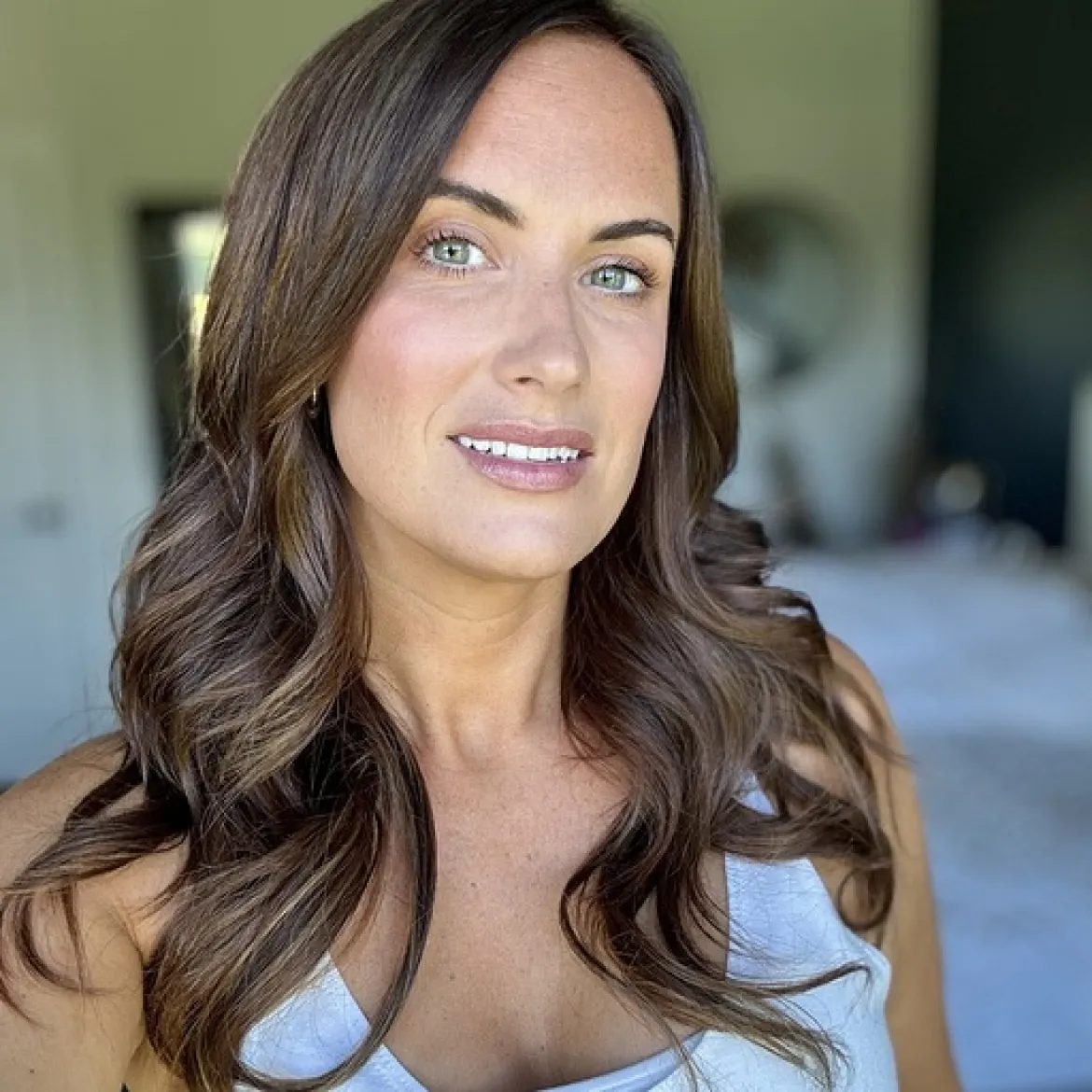 A woman with long brown hair smiling indoors, wearing a light-colored top.