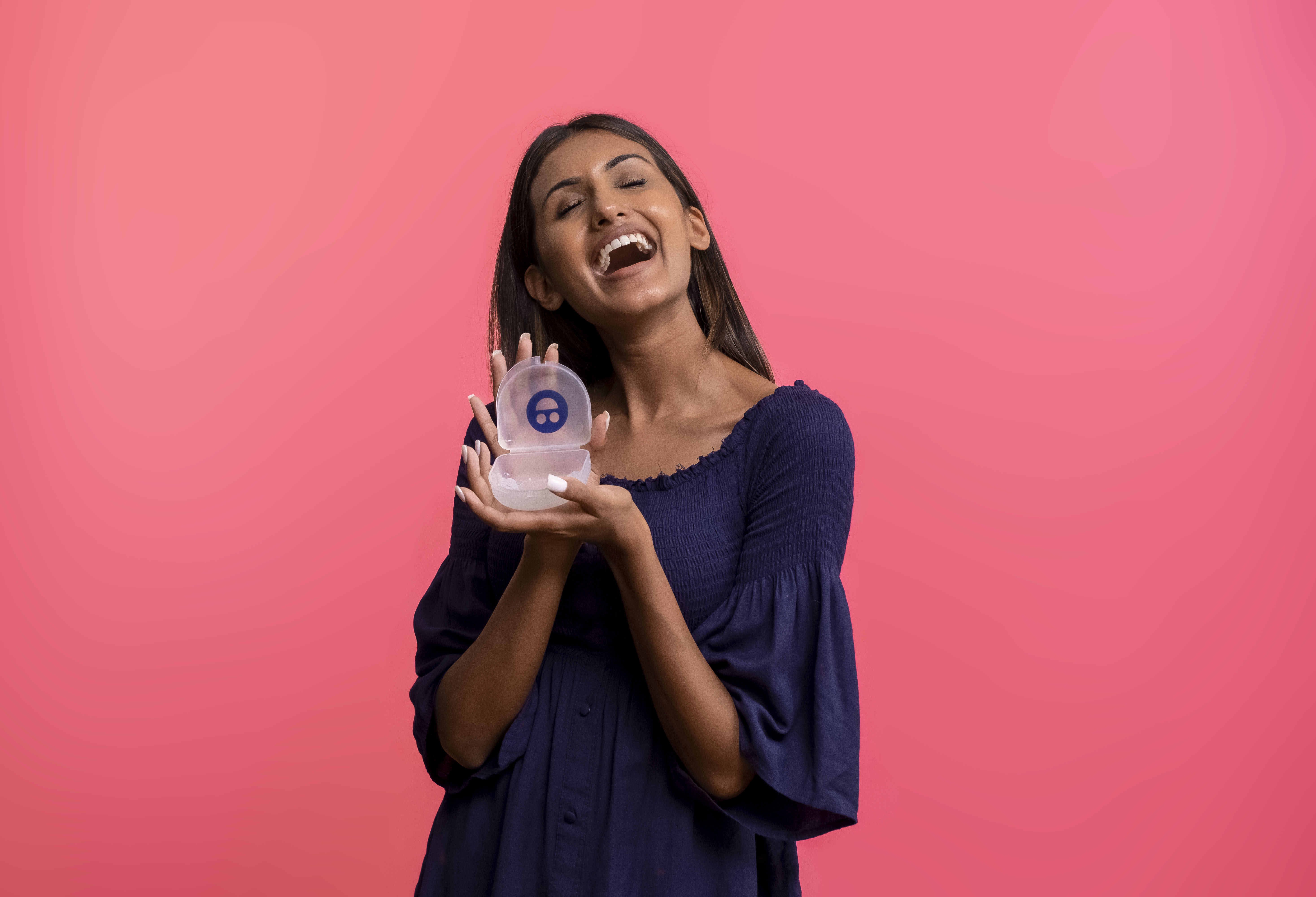Smiling woman holding a small transparent case with a blue symbol, against a pink background.