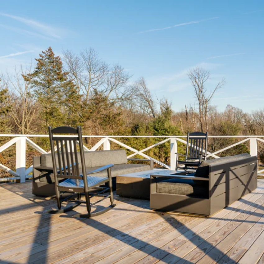 A sunny wooden deck with dark outdoor furniture, including sofas and rocking chairs, overlooking a wooded area.