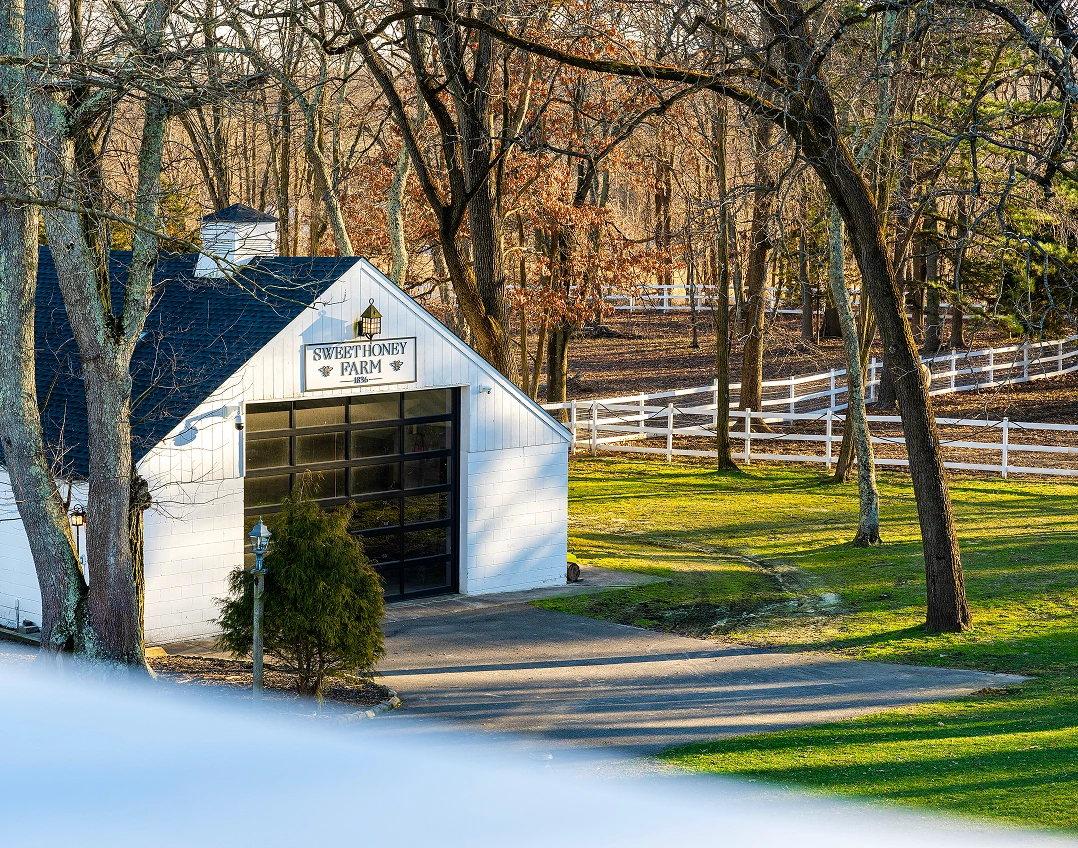 A white barn with a 'Sweet Honey Farm' sign surrounded by bare trees and a white fence.
