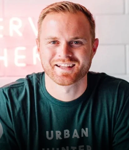 Smiling man with a beard wearing a dark green t-shirt in front of a neon sign.