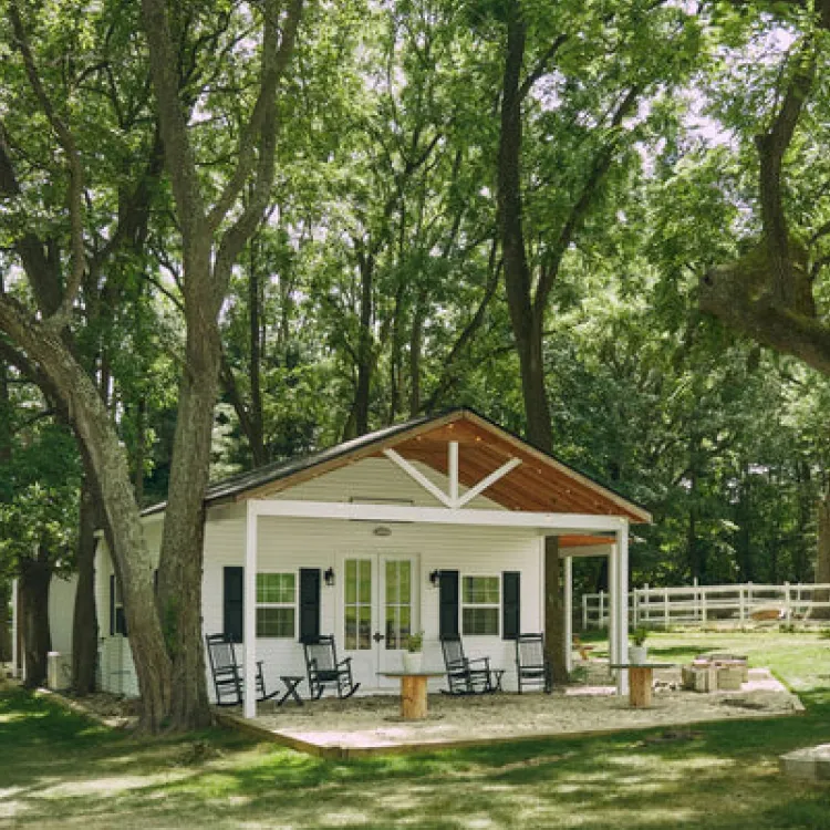 A small white house with a covered porch and rocking chairs, surrounded by tall, leafy trees.