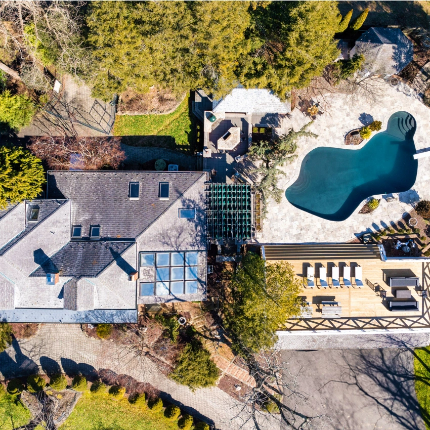 An aerial top-down view of a house with a large, freeform swimming pool, patio, and surrounding trees.
