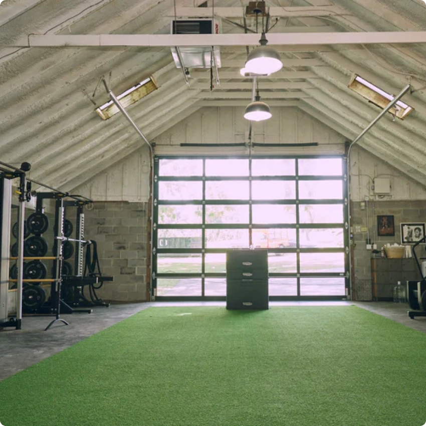 A home gym inside a garage with green turf, weight equipment, and a large, windowed roll-up door.