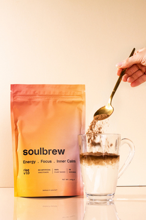 A hand spoons Soulbrew powder into a glass mug of a milky beverage, with the product pouch nearby.