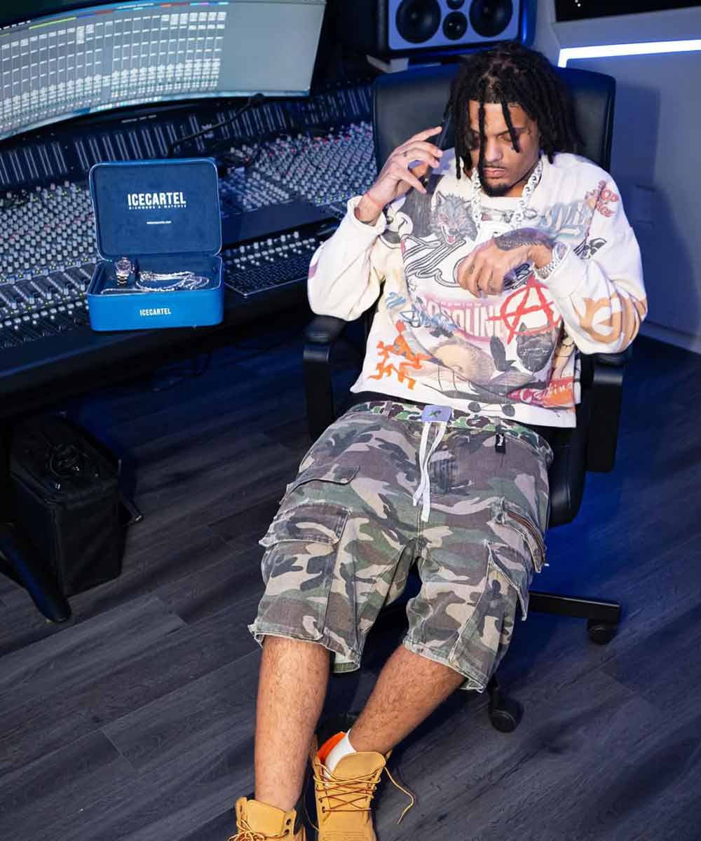 Person sitting in a studio chair, wearing a graphic sweater and camo shorts, with recording equipment nearby.