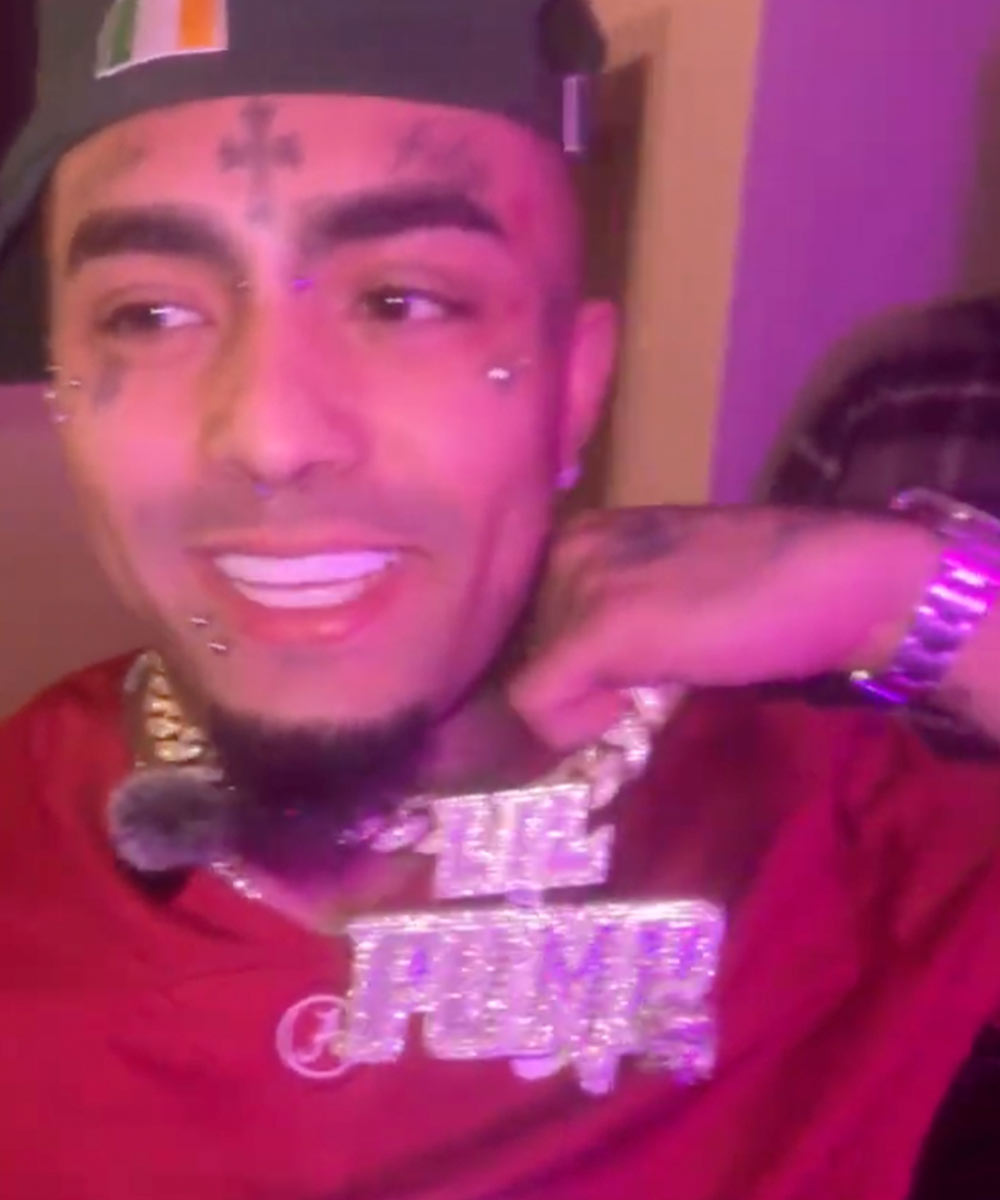 Smiling person with facial tattoos and a large diamond chain.