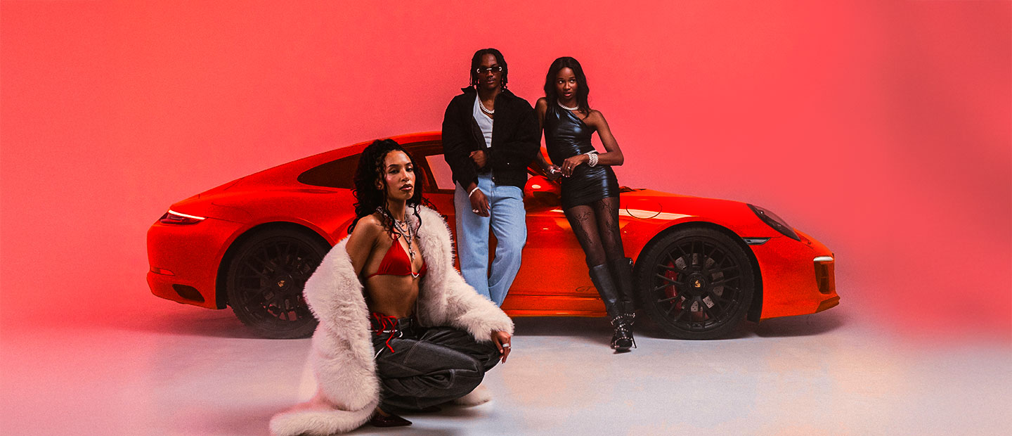 Three people posing with a red sports car against a red background.