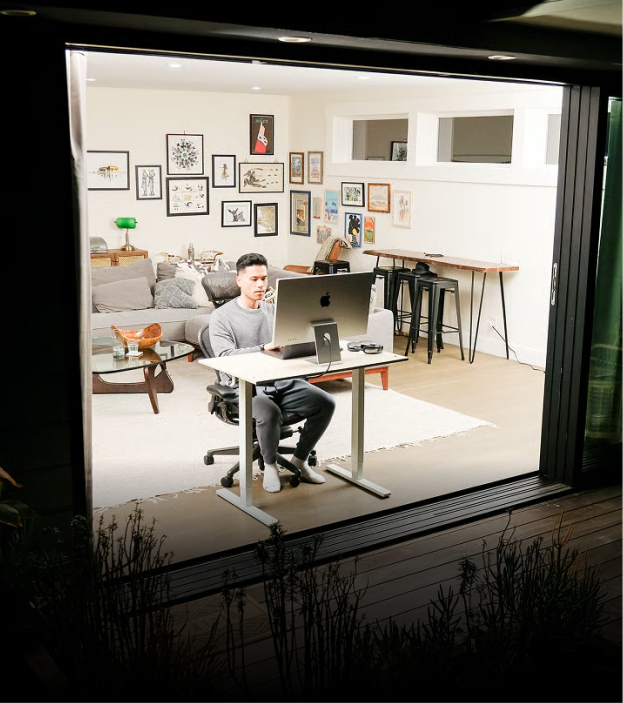 A man works at a standing desk in a brightly lit living room, viewed from outside at night.
