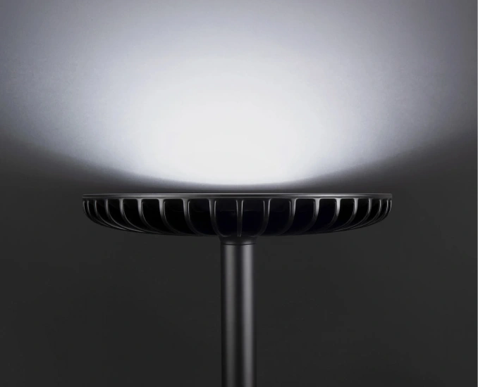 A modern, black, circular lamp with a finned head, illuminating a dark wall with a bright upward light.