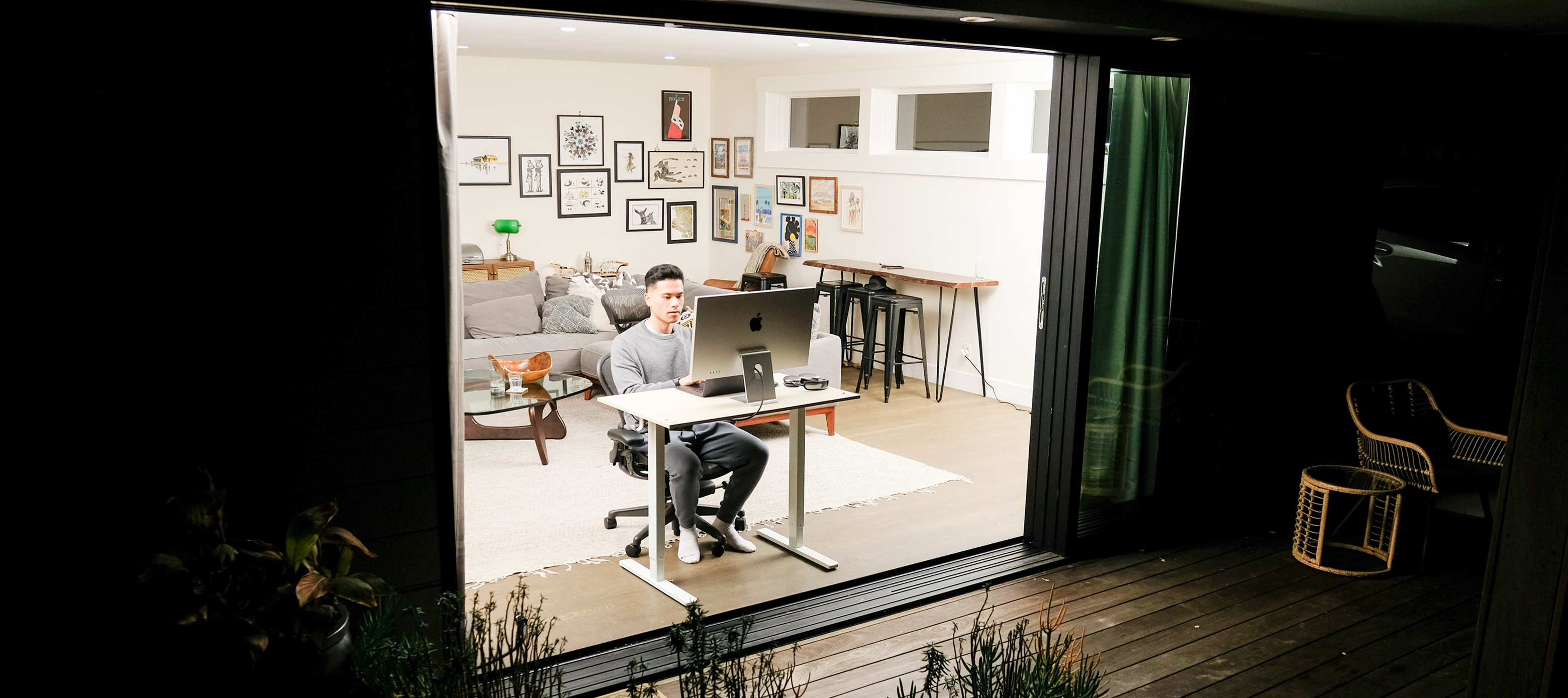 A man works at a desk on a computer in a brightly lit room, viewed from outdoors at night.