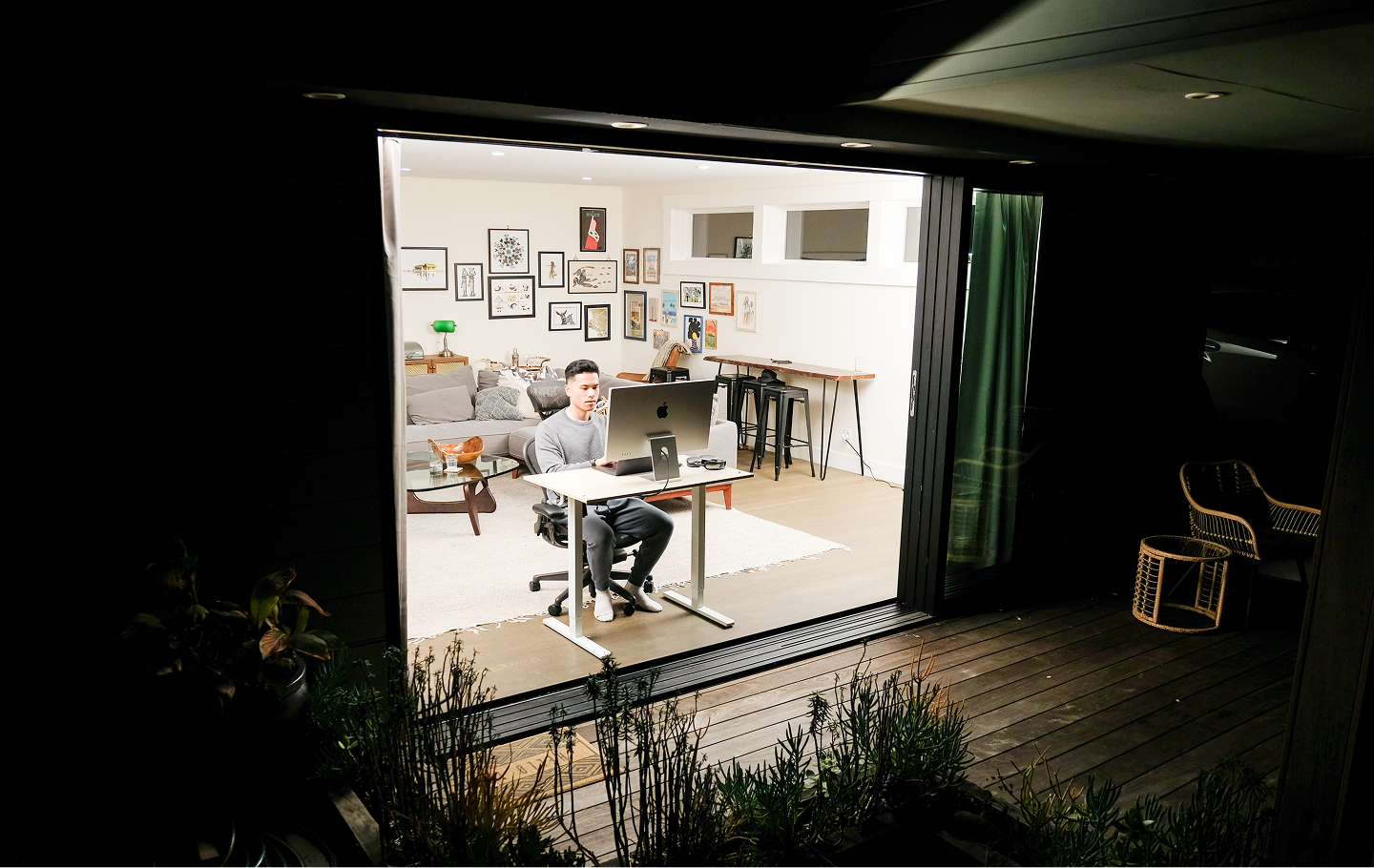 A person works on a computer in a brightly lit room, viewed from a dark patio at night.