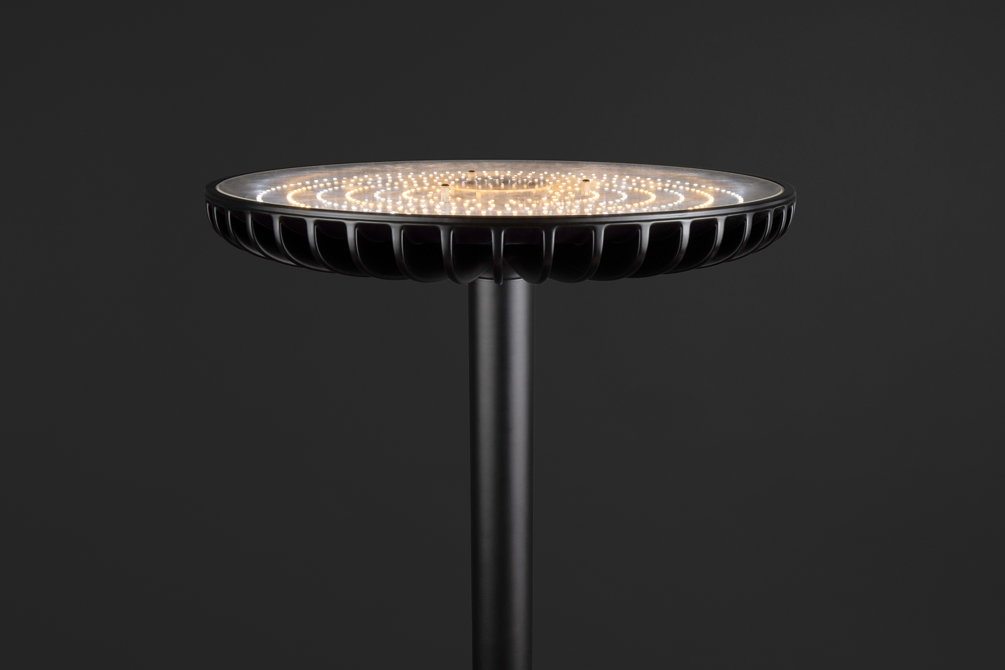 A modern black pole lamp with a circular head of lit LED lights against a dark background.