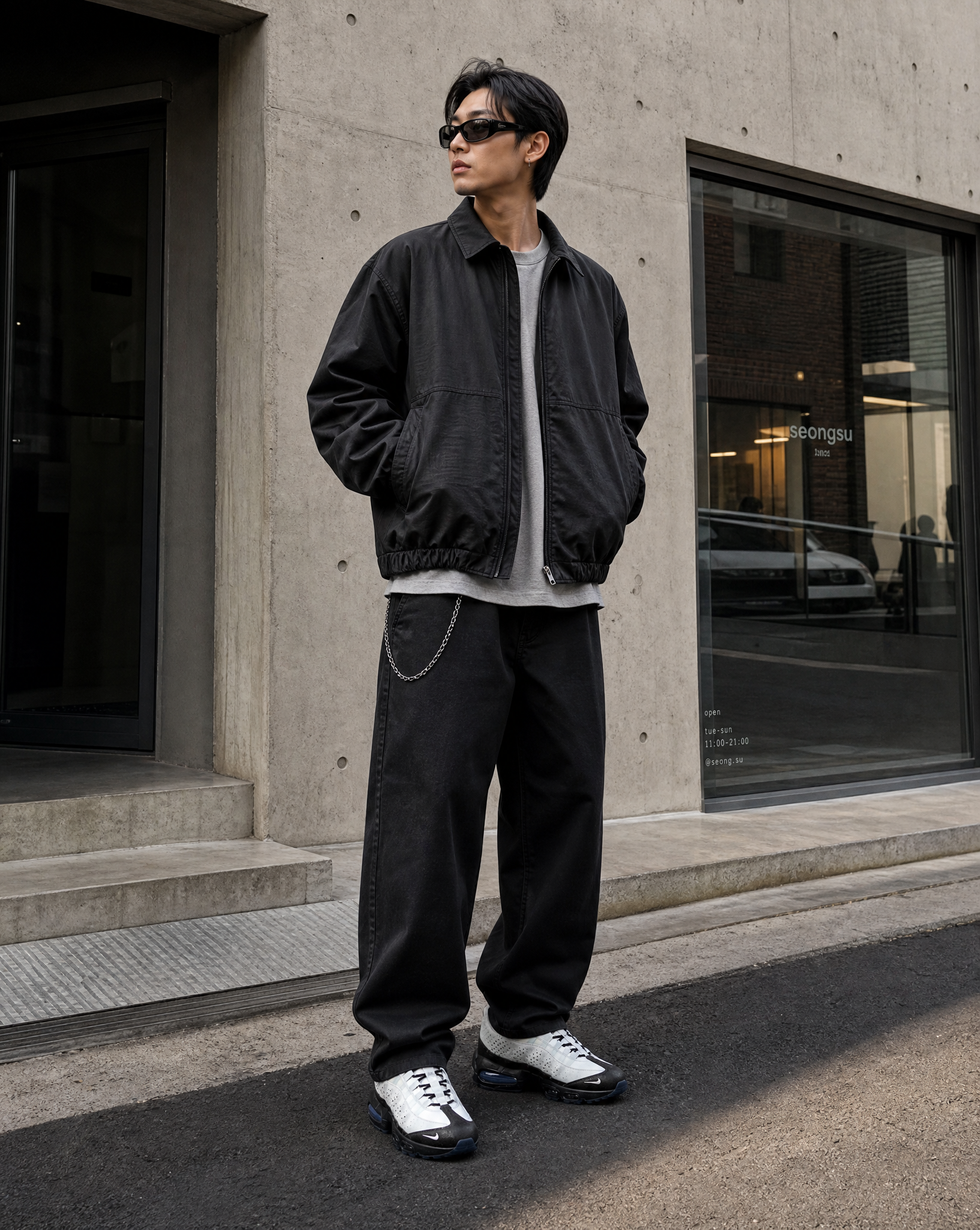 A man in a black jacket, baggy black pants, and sunglasses stands on a city street.