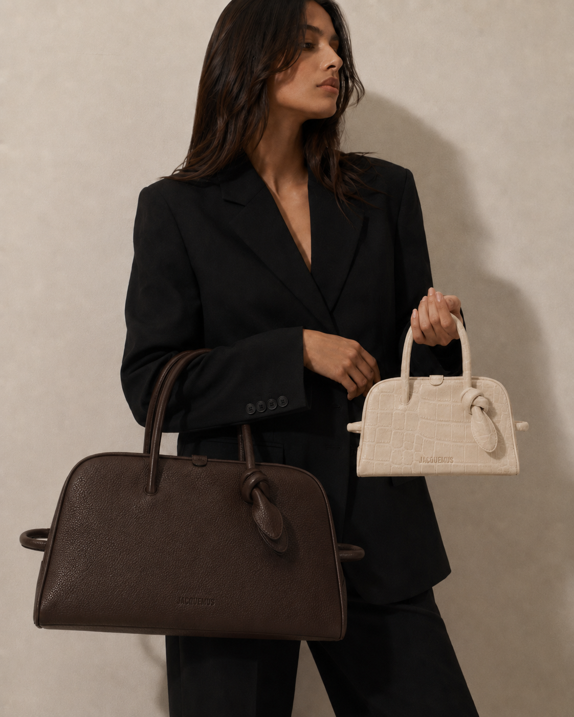 A woman in a black blazer holds two handbags, a large brown one and a smaller cream one.