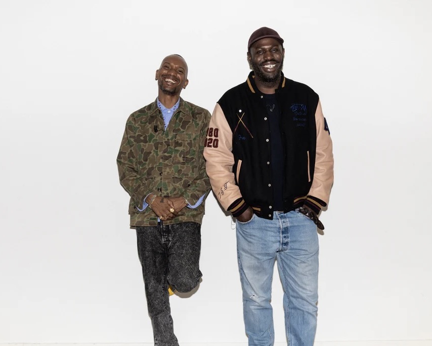Two men smile while standing against a white wall, one wearing a camo jacket, the other a varsity jacket.