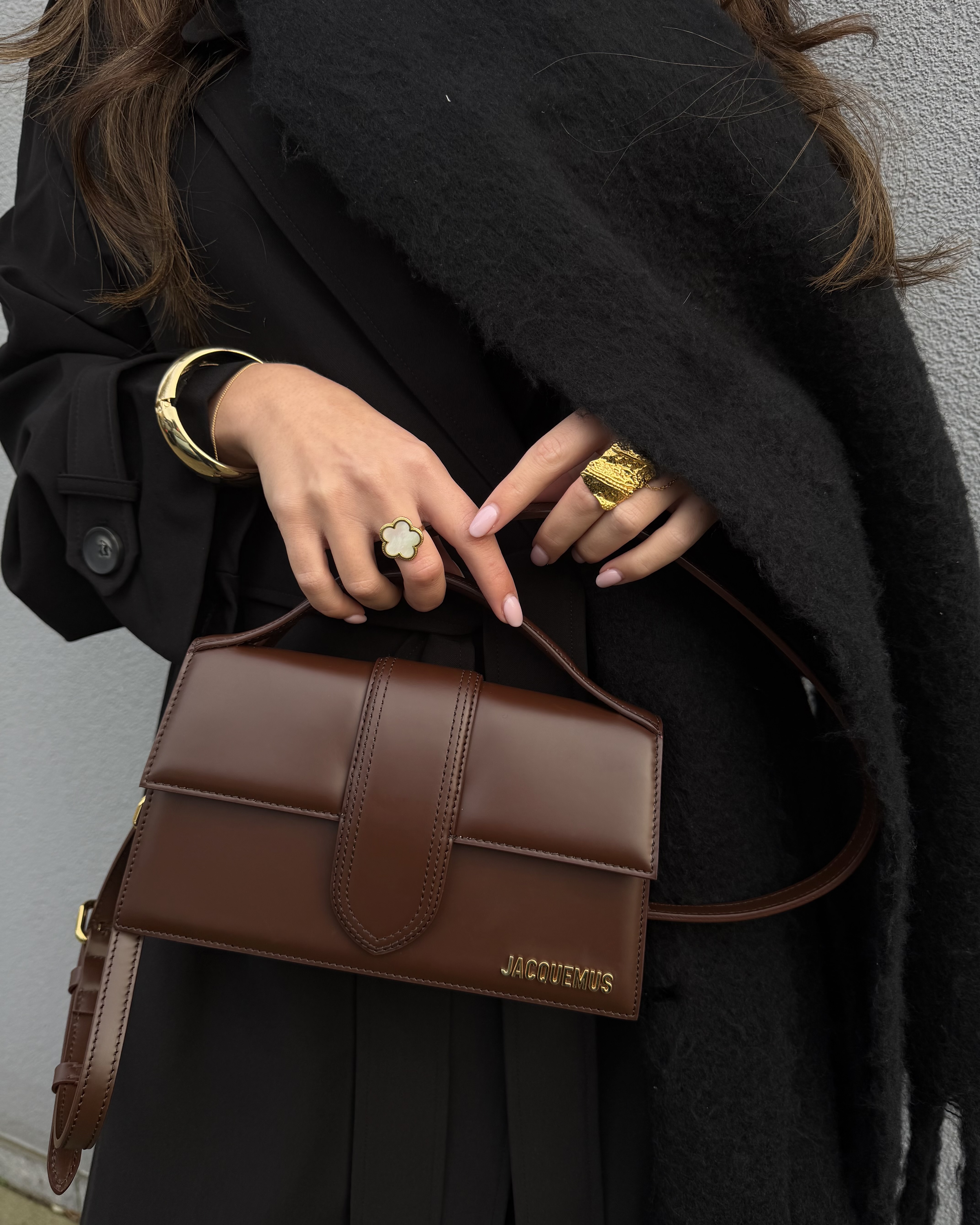 Person holding a brown leather handbag, wearing gold jewelry on a black coat.