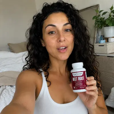 A woman with dark curly hair, wearing a white tank top, holds up a bottle of beet root supplements.