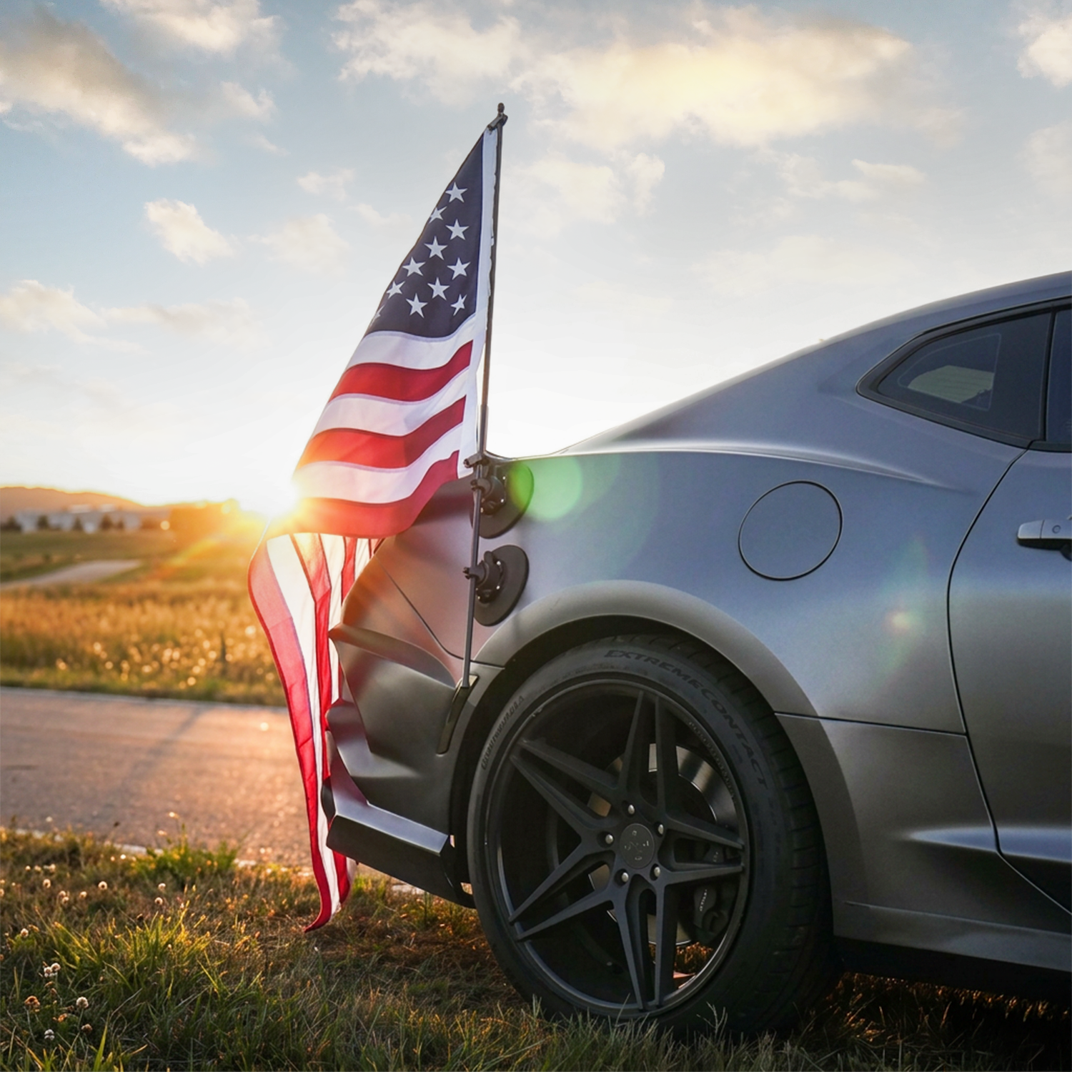 A gray sports car with an American flag mounted on the back, parked on a roadside at sunset.