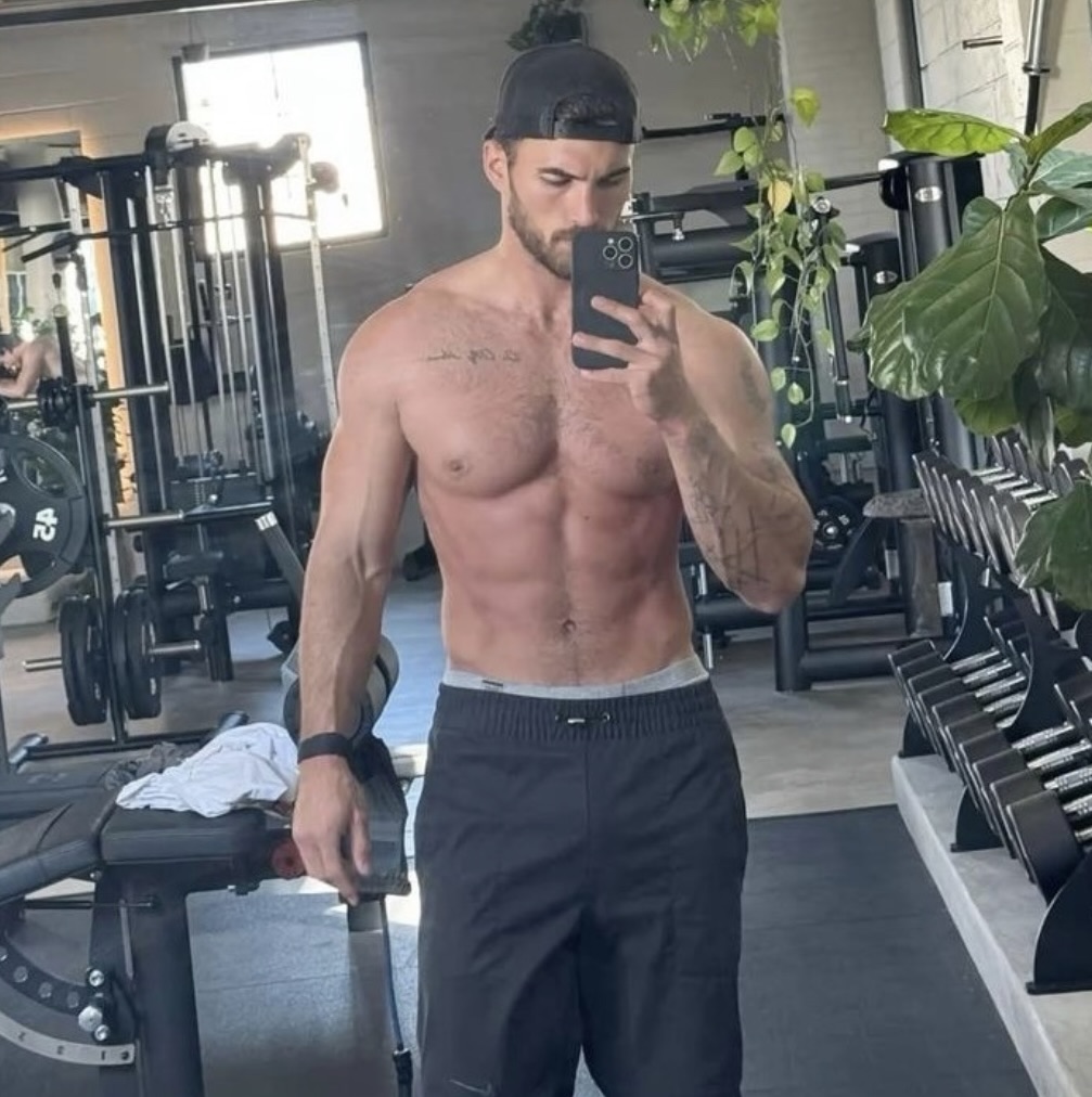 Man taking a mirror selfie in a gym, wearing a cap and workout pants.