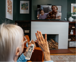 Person watching TV with remote, waving at screen showing two people and a dog.