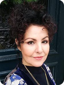 A headshot of a woman with dark, curly hair piled high, smiling in front of a dark door.