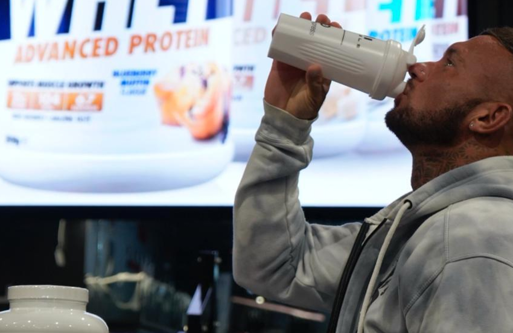 Person drinking from a shaker with protein containers in the background.